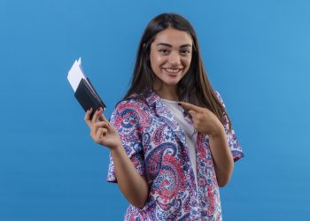 young beautiful woman tourist holding passport with tickets looking at camera pointing with index finger to them smiling cheerfully standing over isolated blue background