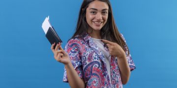young beautiful woman tourist holding passport with tickets looking at camera pointing with index finger to them smiling cheerfully standing over isolated blue background