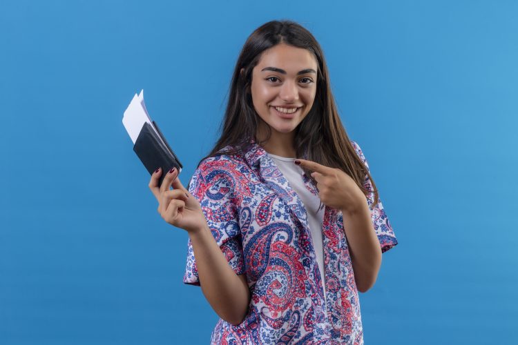 young beautiful woman tourist holding passport with tickets looking at camera pointing with index finger to them smiling cheerfully standing over isolated blue background