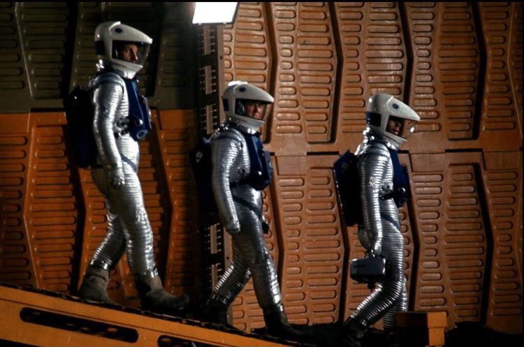 Cena de 2001: Uma Odisseia no Espaço com três astronautas em trajes prateados num ambiente de painéis alaranjados.