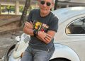 André Giusti, de óculos escuros e braços cruzados, apoia-se em um Fusca branco.