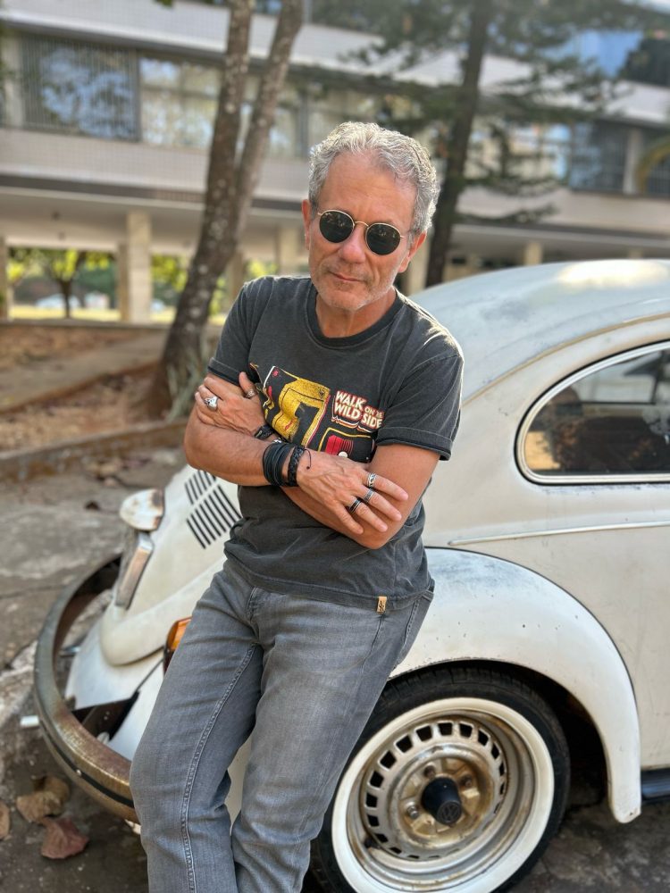 André Giusti, de óculos escuros e braços cruzados, apoia-se em um Fusca branco.