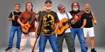 Integrantes da Banda Pra Quem Tá Quente posam com baixo, violão e instrumentos de percussão.