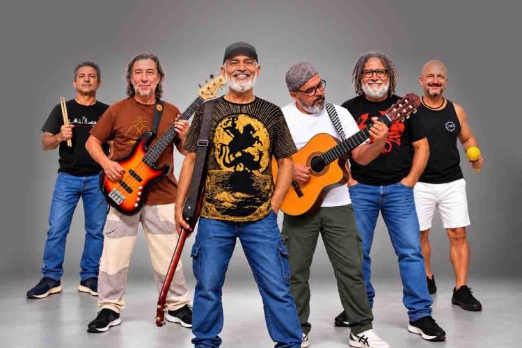 Integrantes da Banda Pra Quem Tá Quente posam com baixo, violão e instrumentos de percussão.