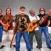 Integrantes da Banda Pra Quem Tá Quente posam com baixo, violão e instrumentos de percussão.