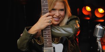 Bia Villa-Chan sorri e abraça uma guitarra preta, diante de luzes alaranjadas e um microfone.