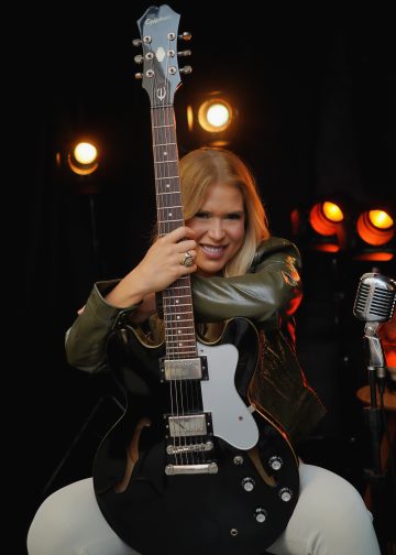 Bia Villa-Chan sorri e abraça uma guitarra preta, diante de luzes alaranjadas e um microfone.