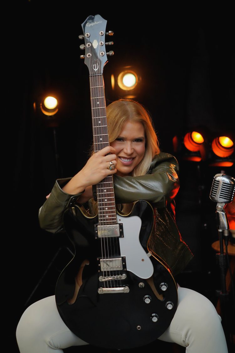 Bia Villa-Chan sorri e abraça uma guitarra preta, diante de luzes alaranjadas e um microfone.