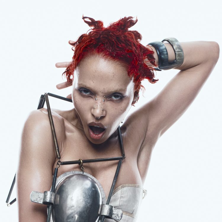 FKA twigs com cabelos vermelhos, maquiagem escura e figurino de peças metálicas em foto de divulgação.