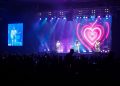 Três integrantes da banda Geminis se apresentam em palco iluminado em azul e rosa, com um coração no telão e público à frente.