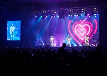 Três integrantes da banda Geminis se apresentam em palco iluminado em azul e rosa, com um coração no telão e público à frente.