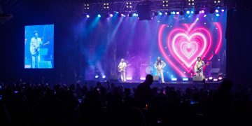 Três integrantes da banda Geminis se apresentam em palco iluminado em azul e rosa, com um coração no telão e público à frente.