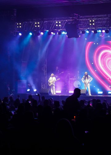 Três integrantes da banda Geminis se apresentam em palco iluminado em azul e rosa, com um coração no telão e público à frente.