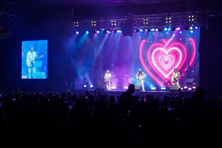 Três integrantes da banda Geminis se apresentam em palco iluminado em azul e rosa, com um coração no telão e público à frente.