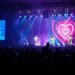 Três integrantes da banda Geminis se apresentam em palco iluminado em azul e rosa, com um coração no telão e público à frente.