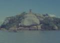 Imagem da Ilha do Presídio, no Guaíba, com rochas, vegetação e uma construção ao fundo.