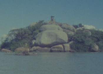 Imagem da Ilha do Presídio, no Guaíba, com rochas, vegetação e uma construção ao fundo.