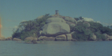 Imagem da Ilha do Presídio, no Guaíba, com rochas, vegetação e uma construção ao fundo.