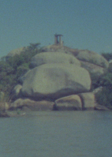 Imagem da Ilha do Presídio, no Guaíba, com rochas, vegetação e uma construção ao fundo.