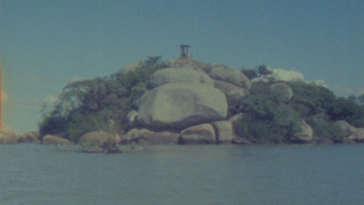 Imagem da Ilha do Presídio, no Guaíba, com rochas, vegetação e uma construção ao fundo.