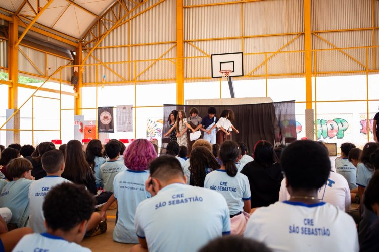 Estudantes apresentam uma cena diante da comunidade escolar, em uma quadra coberta, no projeto de leitura dramatizada no DF.