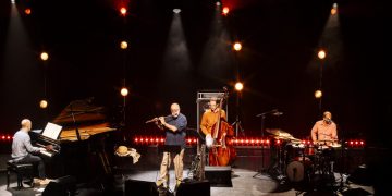 Teco Cardoso Quarteto no palco, com flauta, piano, contrabaixo e bateria, sob iluminação em tons quentes.