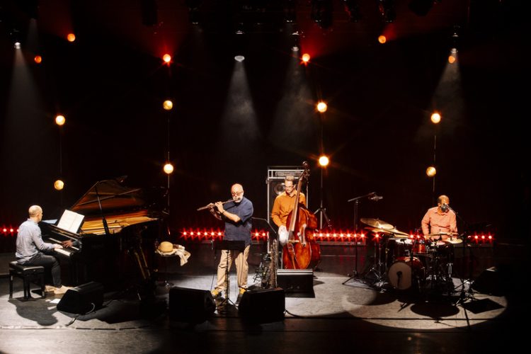 Teco Cardoso Quarteto no palco, com flauta, piano, contrabaixo e bateria, sob iluminação em tons quentes.
