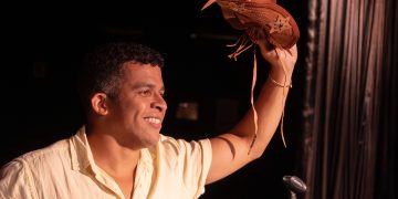 Wêlson Nunes sorri no palco, com um microfone na mão e um chapéu de couro erguido.