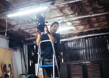 Zizi Antunes ajusta equipamento de iluminação cênica em uma escada durante atividade técnica