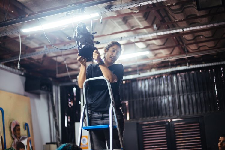 Zizi Antunes ajusta equipamento de iluminação cênica em uma escada durante atividade técnica